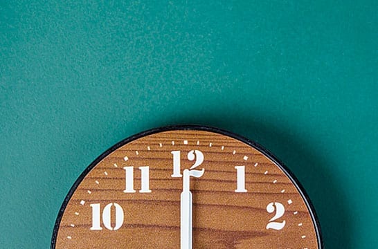 Wood clock with hands at noon against a green wall